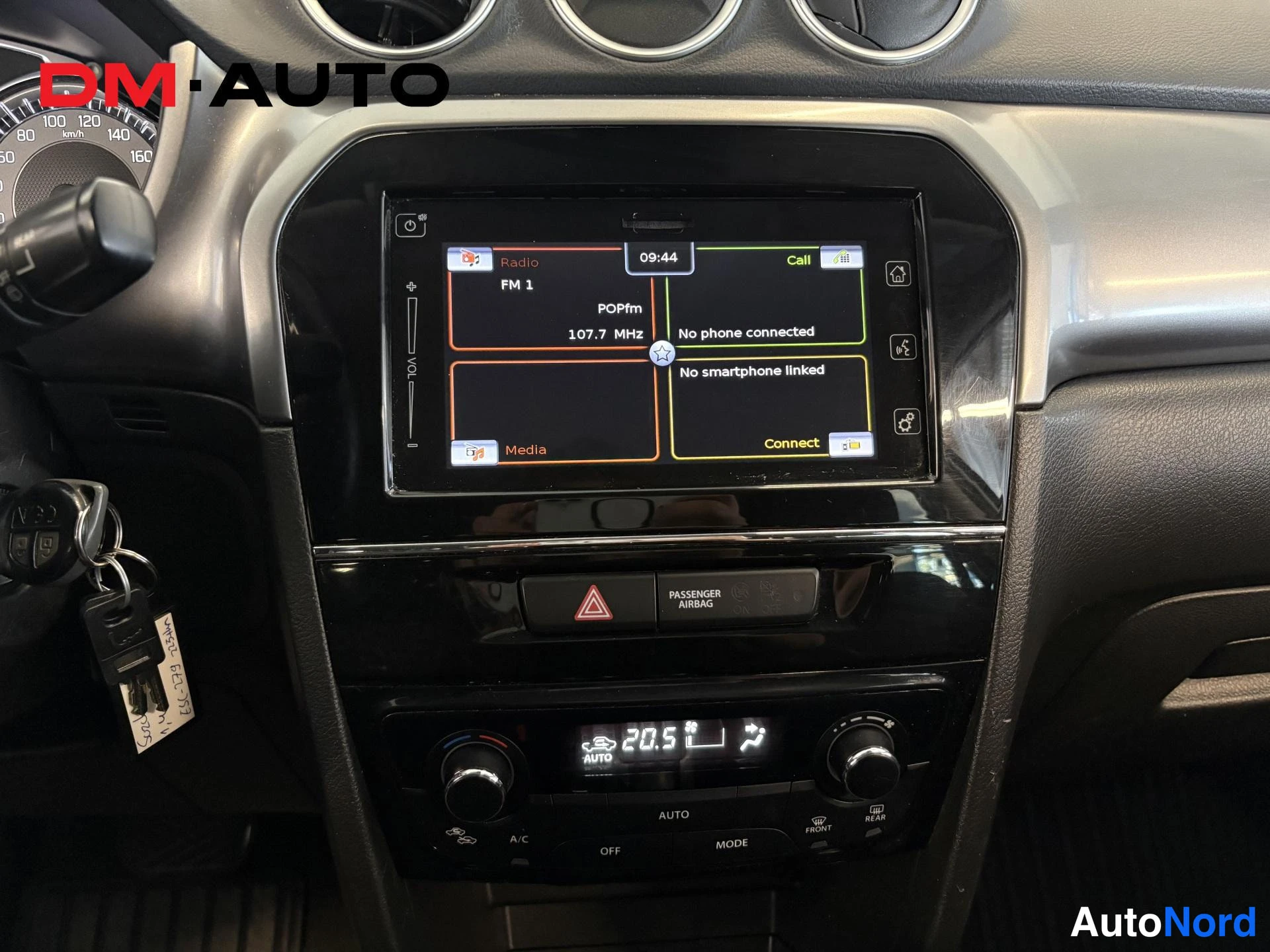 Etuviisto: Keskikonsolin kosketusnäyttö radiolla (FM 1, POPfm 107.7 MHz) ja tekstillä 'No phone connected' ja 'No smartphone linked'; alla varoituskolmiopainike, matkustaj