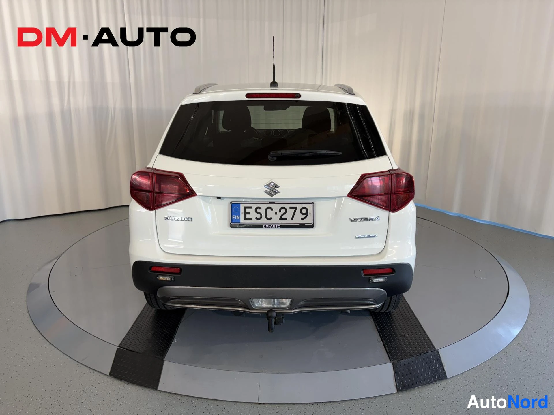 Takaa: Valkoisen tila-auton takaosa suoraan edestä, rekisterikilpi, Suzuki- ja Vitara-merkit, vetokoulu.