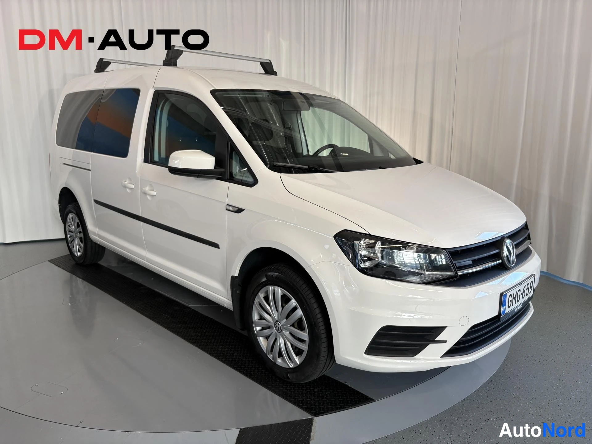 Etuviisto, nokka oikealle: Valkoinen Volkswagen Caddy etuosakolmannes oikealta kuvattuna hallissa, katolla kattotelineet.
