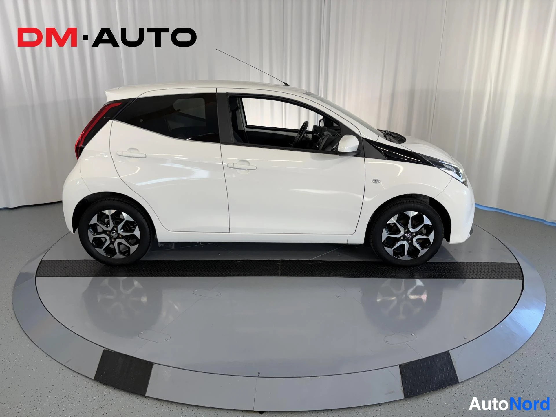 Sivulta: Valkoinen Toyota Aygo -henkilöauto sivusta vasemmalta, mustat kevytmetallivanteet, kuljettajan oven auki.