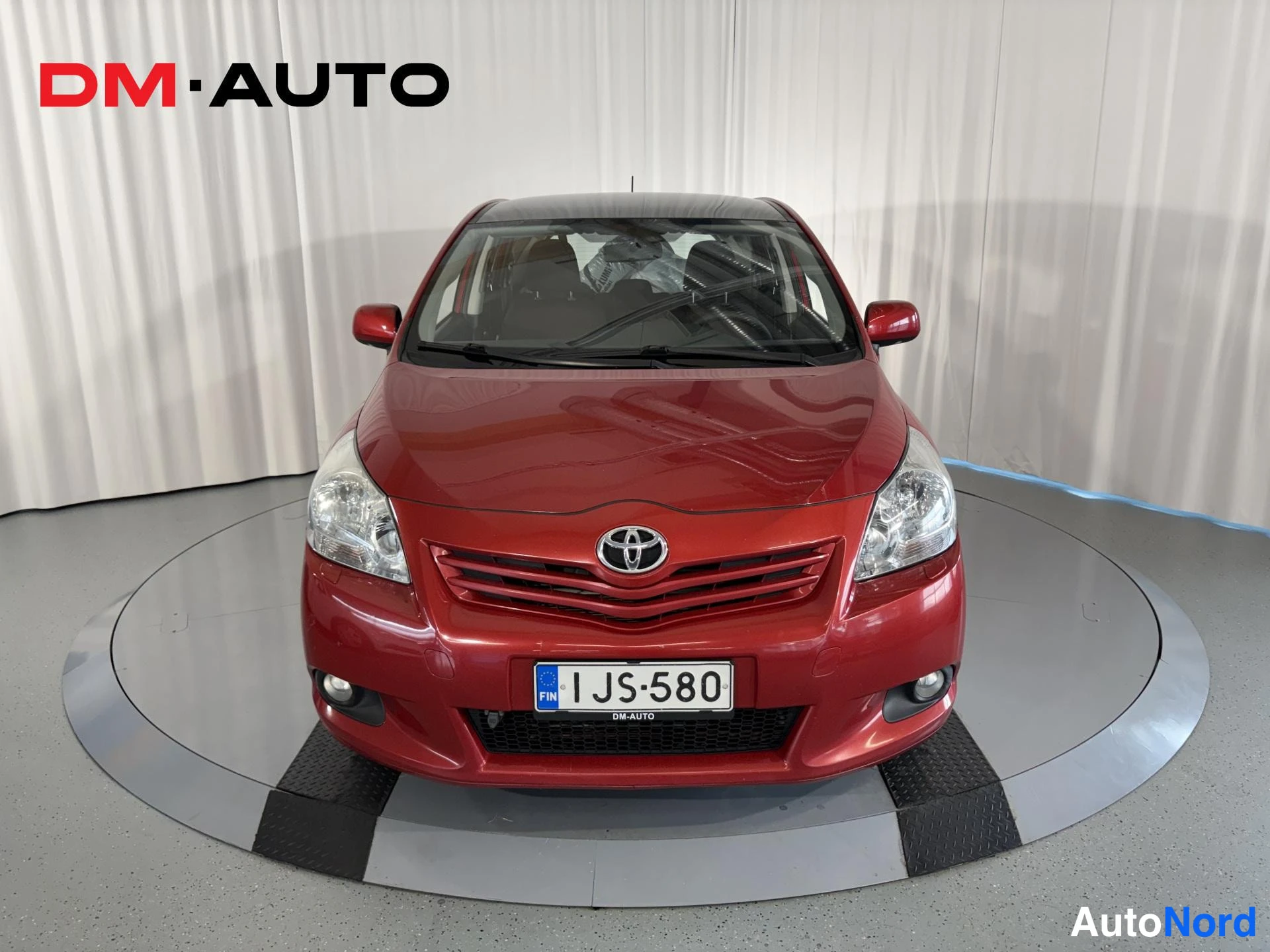 Edestä: Punaisen Toyota Verso -tilavaunun etunäkymä suoraan edestä valkotaustaa vasten; rekisterikilpi näkyvissä.