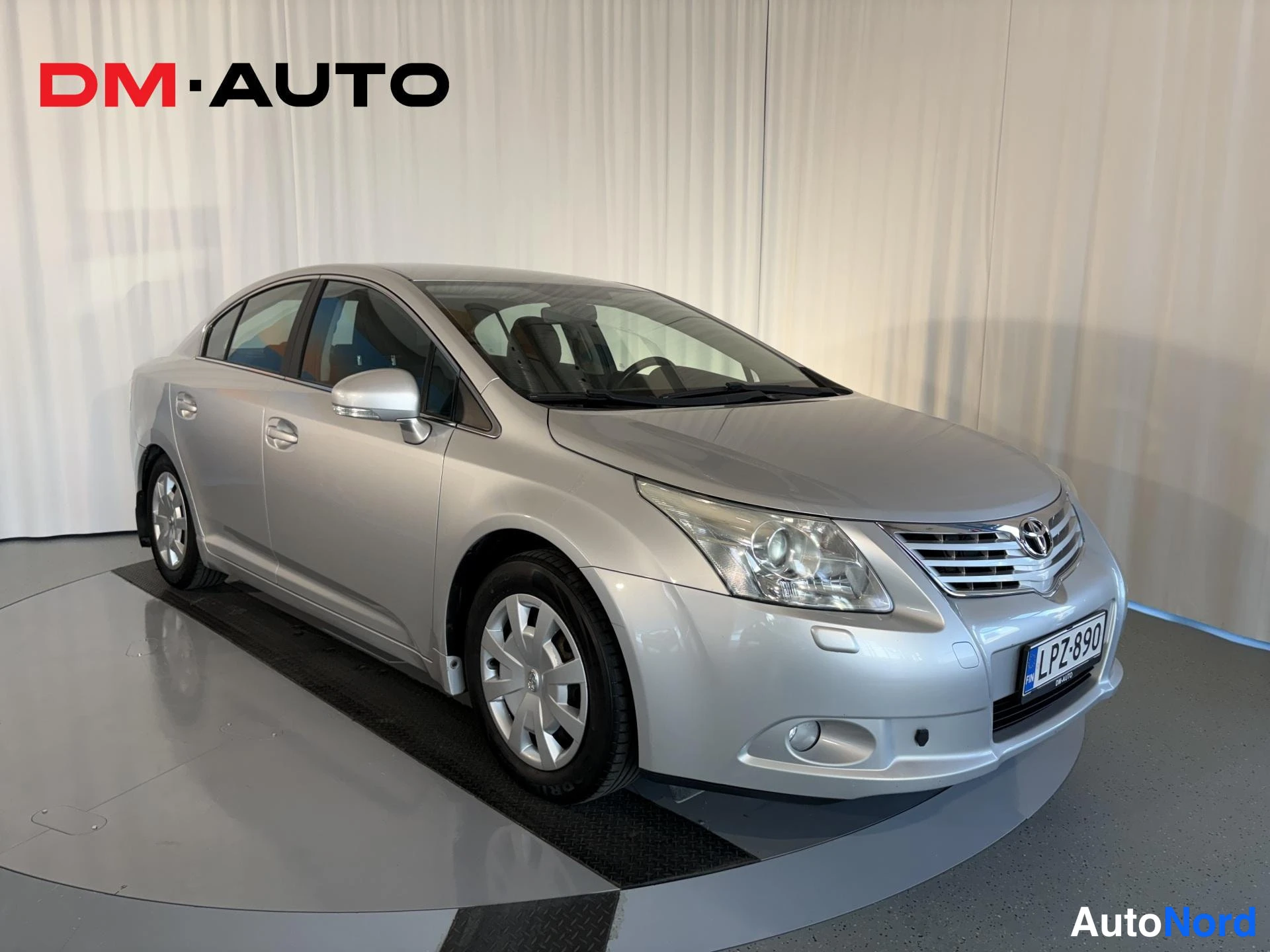 Etuviisto, nokka oikealle: Toyota Avensis -sedanin etuosa kolmivartalokuvassa käänteisestä kulmasta, hopeinen kori ja rekisterikilpi