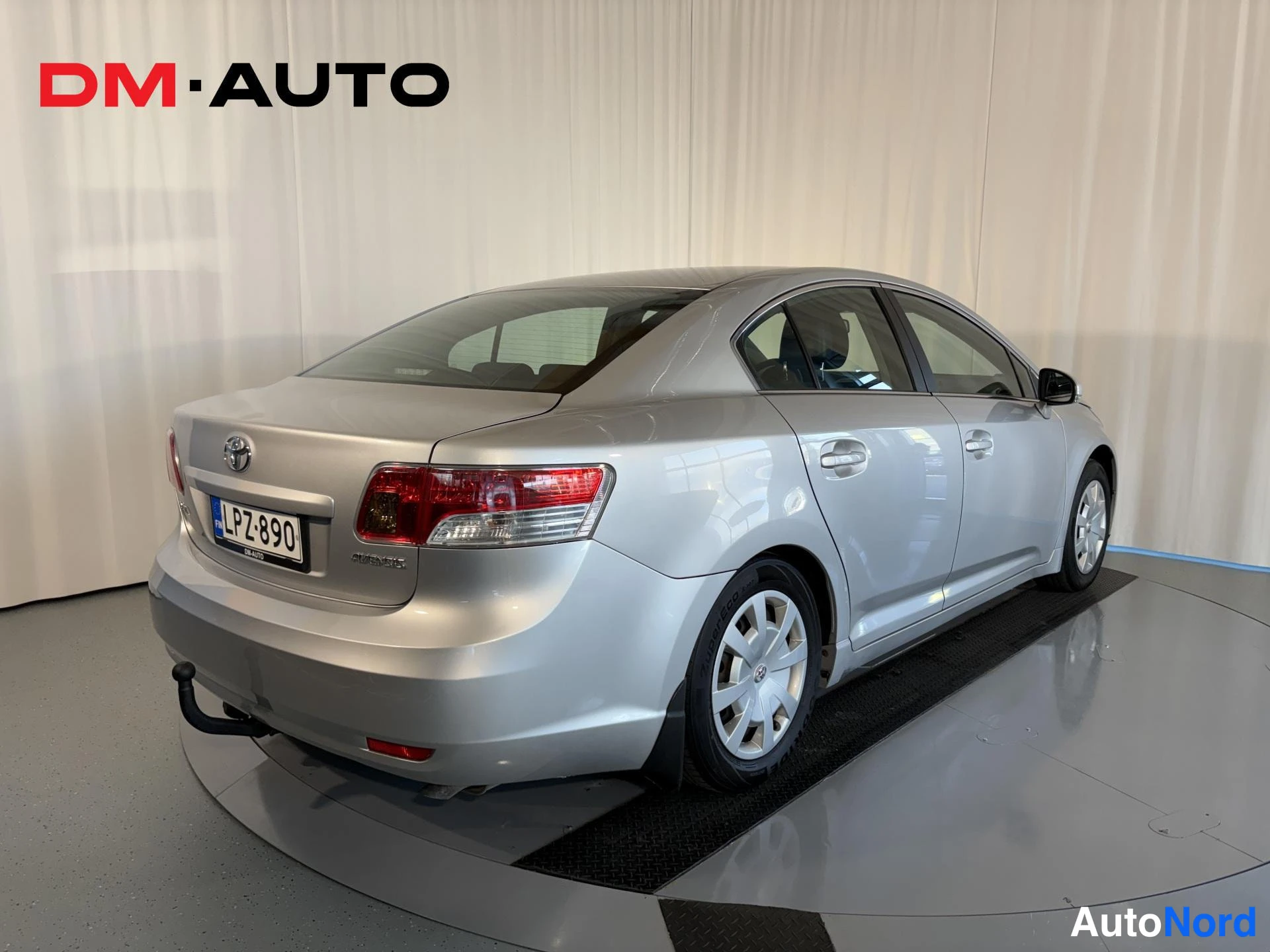 Takaviisto: Toyota Avensis -sedanin takakulmanäkymä: hopeainen kori, takavalot ja rekisterikilpi näkyvät valokuitutaustaa vasten.