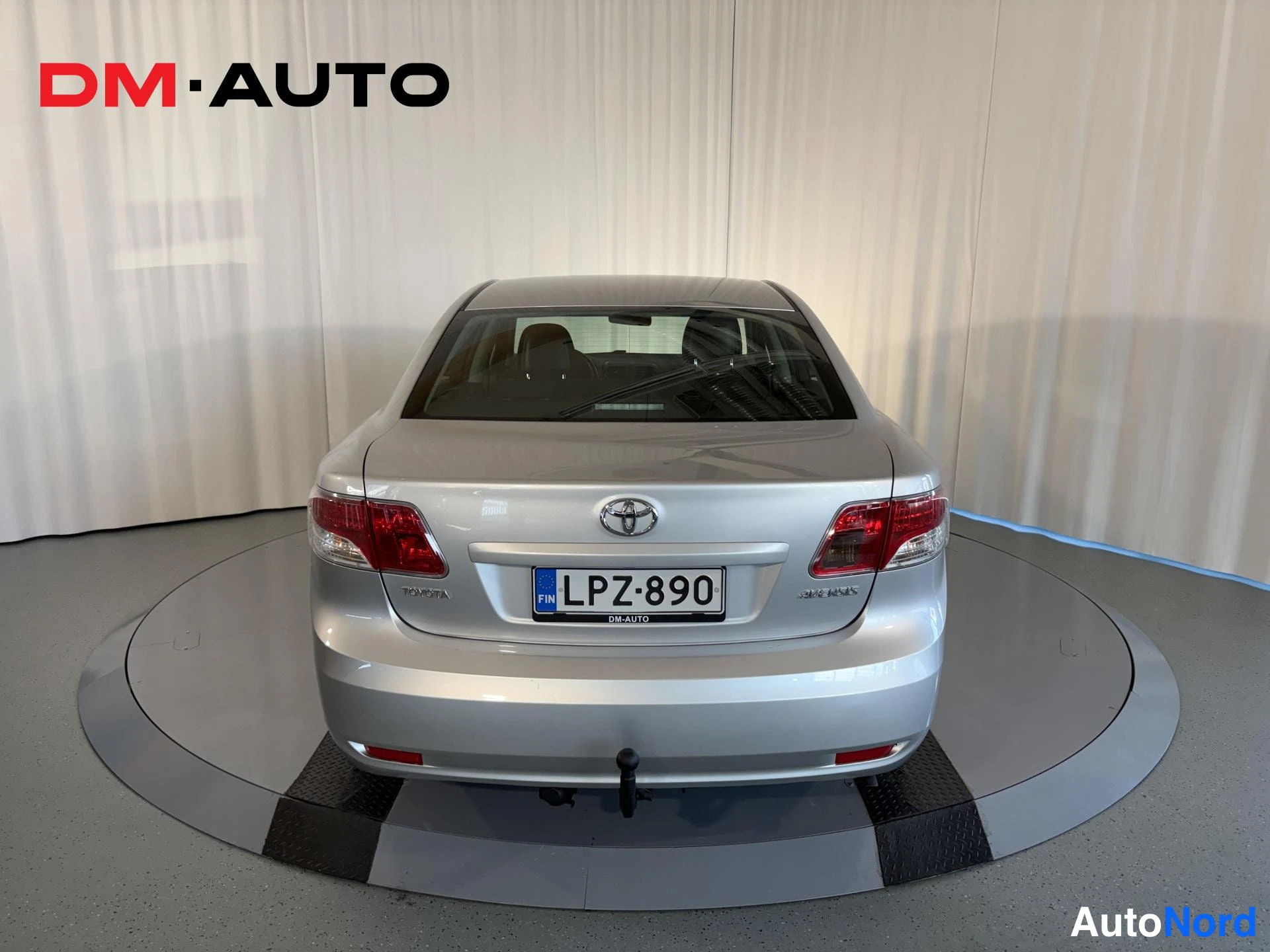 Takaa: Toyota Avensis -sedanin suora taka-näkymä: Toyota-merkki, punaiset takavalot, rekisterikilpi ja tahdistettu perävaunun kytkin.