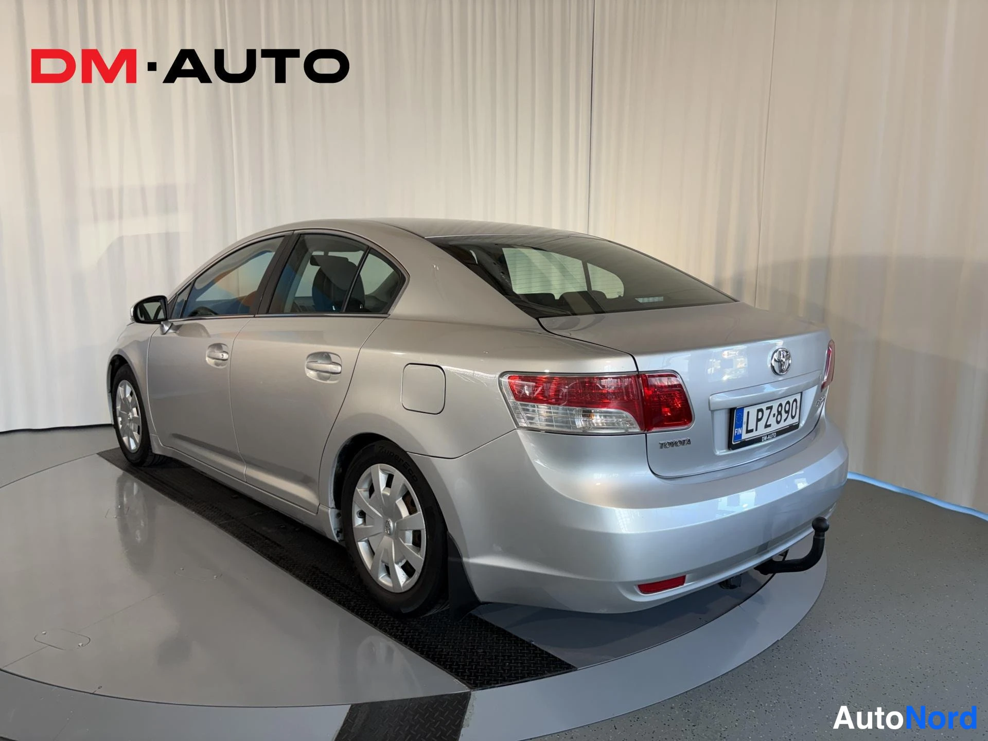 Takaviisto: Toyota Avensis -sedanin takakulmanäkymä vasemmalta: hopeinen kori, takavalot, rekisterikilpi ja poistuva perävaunun kytkin.