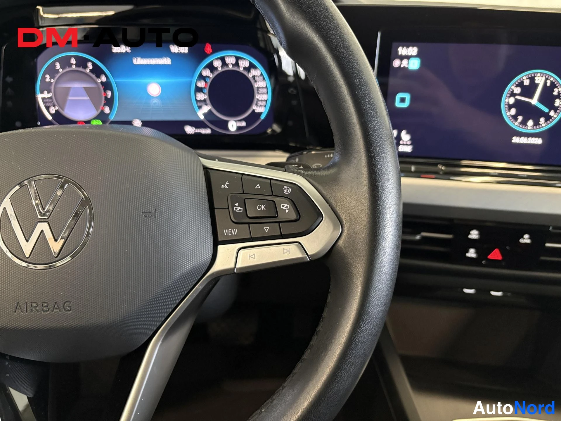 Edestä: Rattipychy Volkswagen-logolla ja AIRBAG-tekstillä, taustalla digitaalinen mittaristo ja kosketusnäyttö.