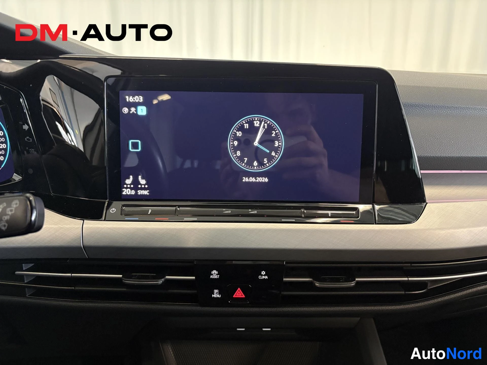 Edestä: Volkswagenin kojelaudan keskialue: kosketusnäyttö kellon ja 26.06.2026 päivämäärän kanssa, alapuolella ilmastointipaneeli.