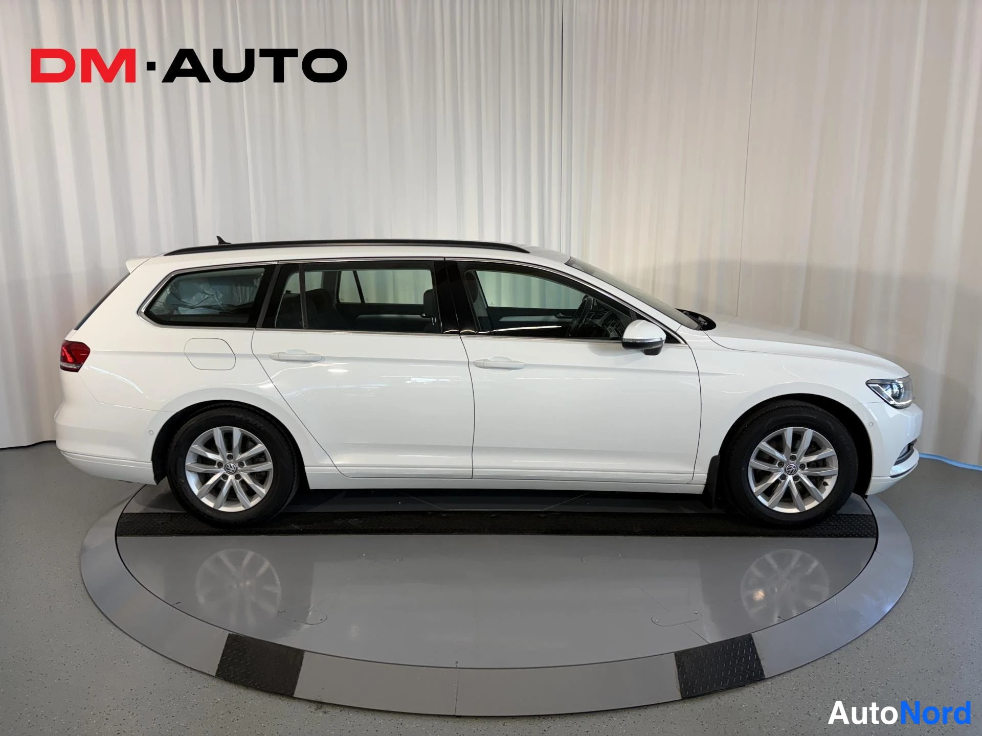Sivulta: Valkoinen Volkswagen Passat Variant sivulta kuvattuna, koko kylki ja pyörät näkyvät, kattokaiteet, LE-divaani, taustavalot reunassa.
