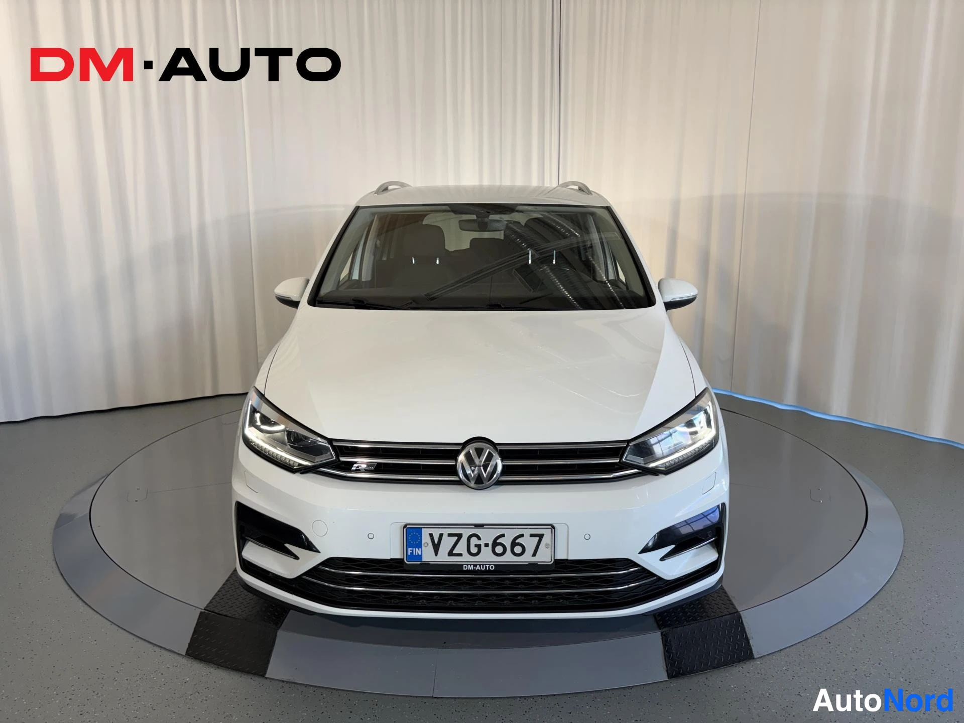 Edestä: Valkoisen tila-auton suora etukuva; etusäleikkö ja Volkswagen-merkki näkyvät, rekisteritunnus.