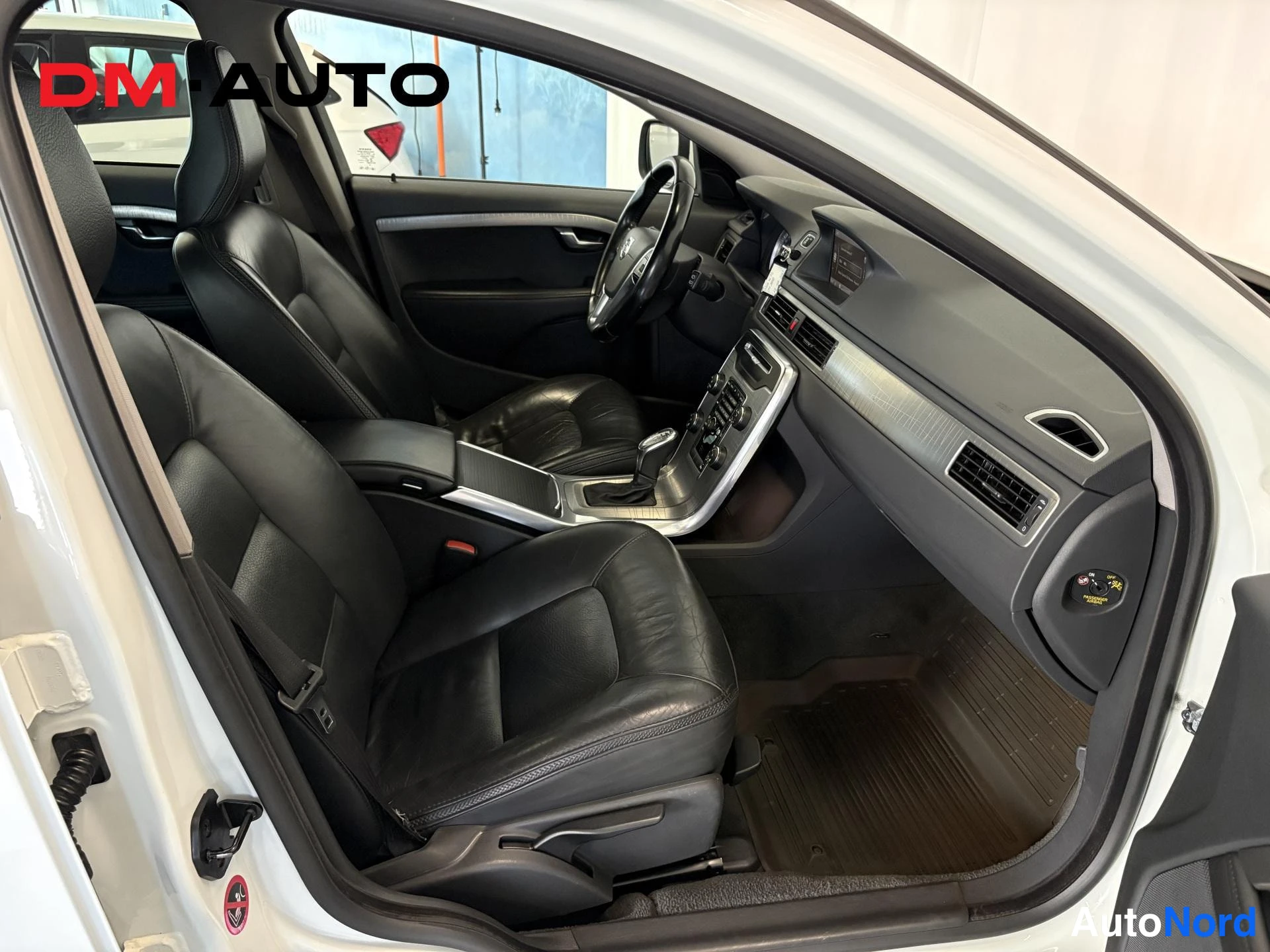 Muu: Autojen sisätilat: mustat nahkaistuimet, ohjauspyörä ja kojelauta kuvattuna avoimesta etuovesta.