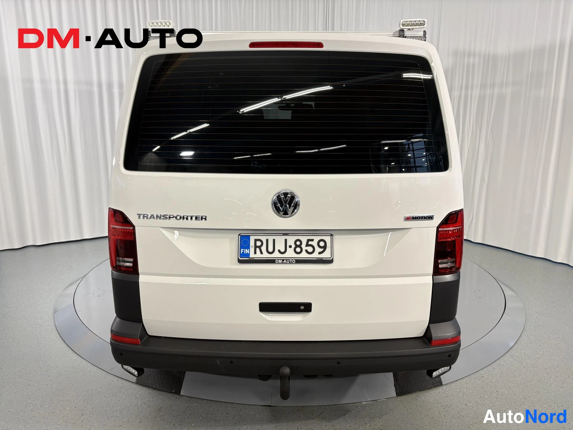 Takaa: Valkoisen pakettiauton takaosa suoraan edestä: TRANSPORTER-teksti, VW-merkki, rekisterikilpi ja perävaunukoukku.