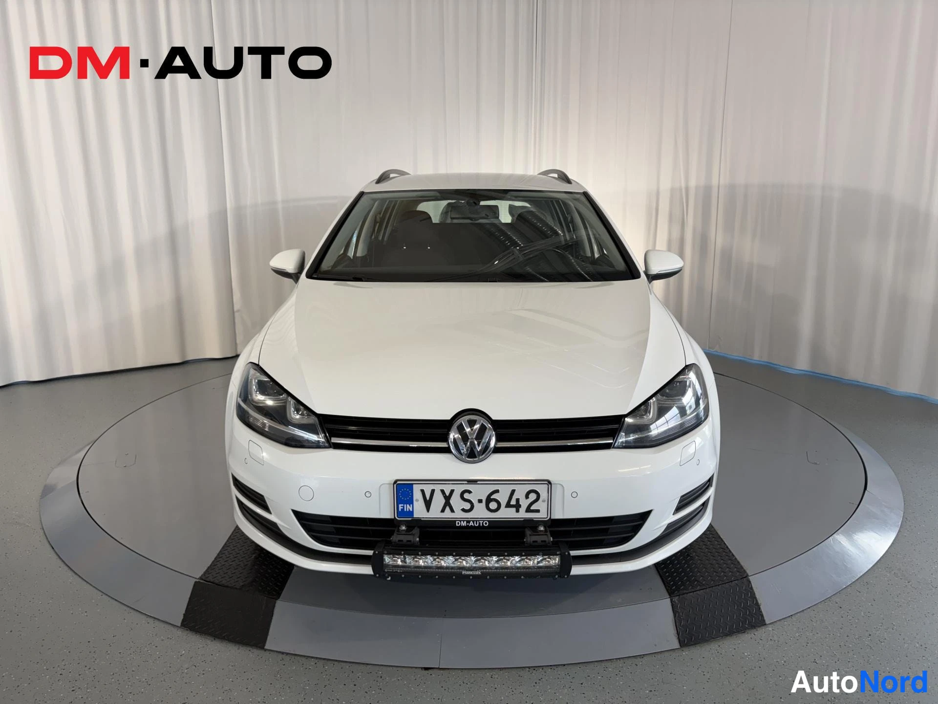 Edestä: Valkoinen farmariauto suoraan edestä, etusäleikkö ja Volkswagen-merkki näkyvissä, rekisterikilpi ja lisävalo puskurissa.