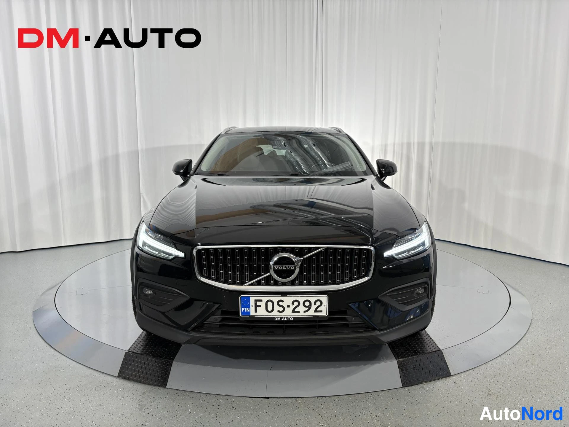 Edestä: Mustan farmariajan Volvo suoraan edestä valokuvattuna, rekisterikilpi ja DM-Auto-logo taustalla.