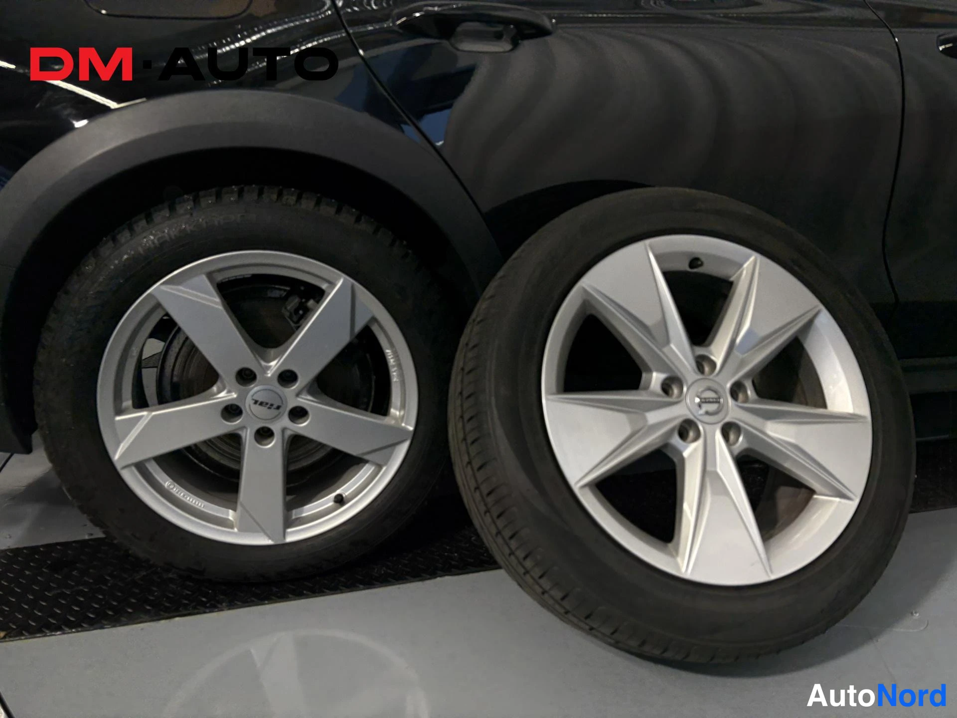 Muu: Mustan auton takapyörä ja vieressä lattialla seistävä Volvo-kekavara, kaksi hopeavannelle.