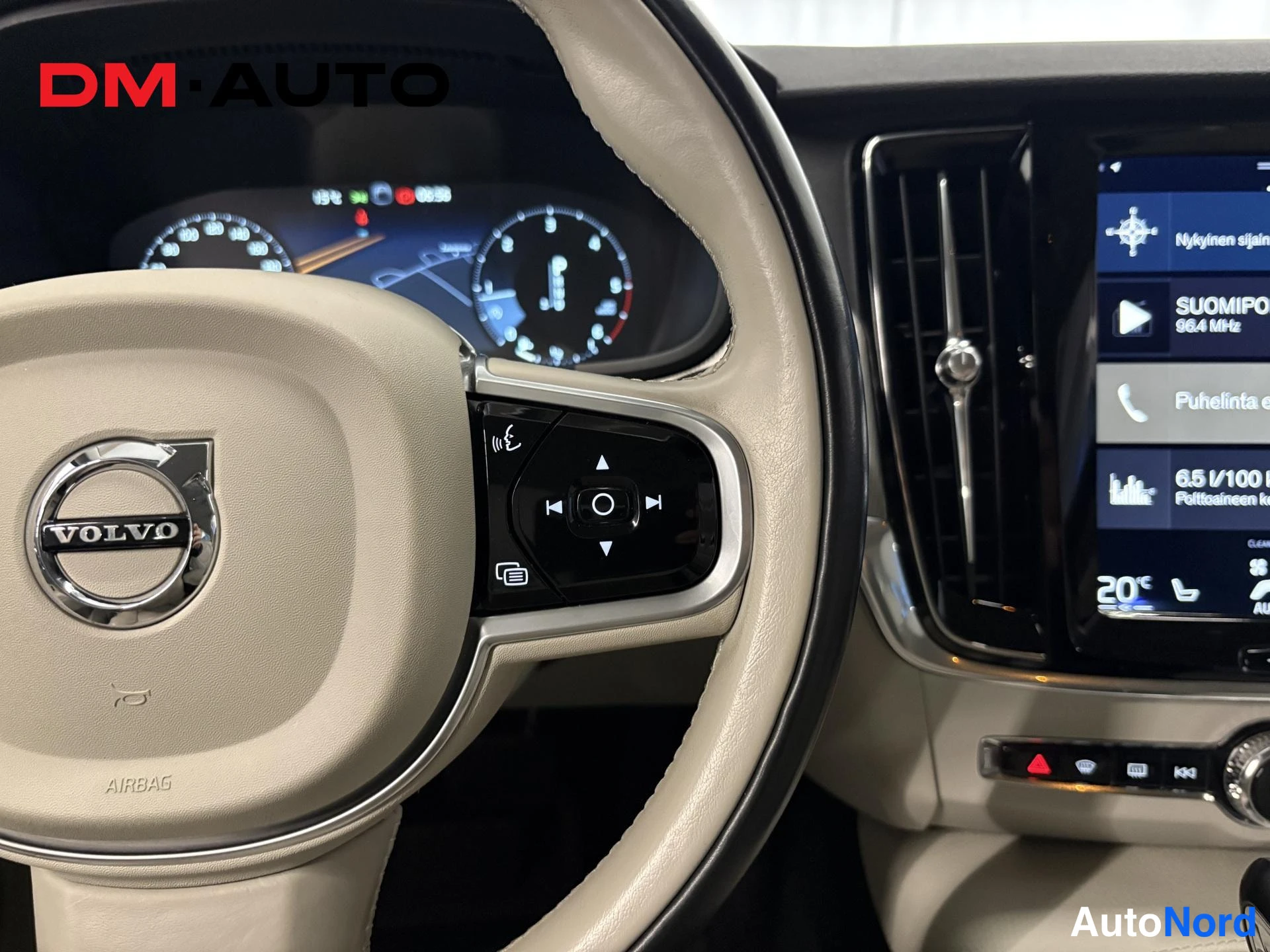 Muu: Ohjauspyörän ja kojelaudan lähikuva: beige Volvo-ohjauspyörä, digitaalinen mittaristo ja kesk Näytön vasen osa radio- ja puhelinvalikoineen.
