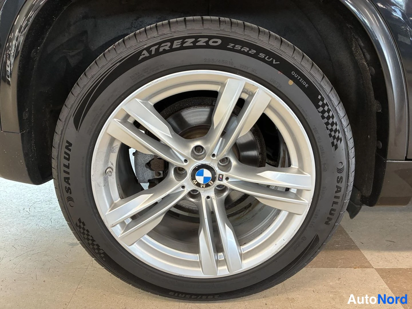 Muu: Lähikuva auton pyörästä: hopea BMW-kehysalusvvana ja musta Sailun Atrezzo 235/55 R19 -rengas, jarrulevy näkyvissä vanteen raodista.