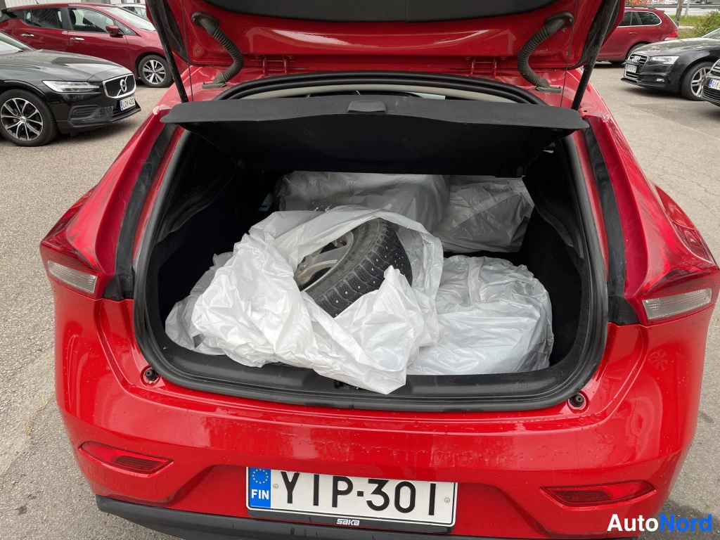 Takaa: Auki avattu tavaratila punaisessa autossa; tilassa renkaat ja lamppuja suojaavat valkoiset muovipussit, rekisterikilpi.