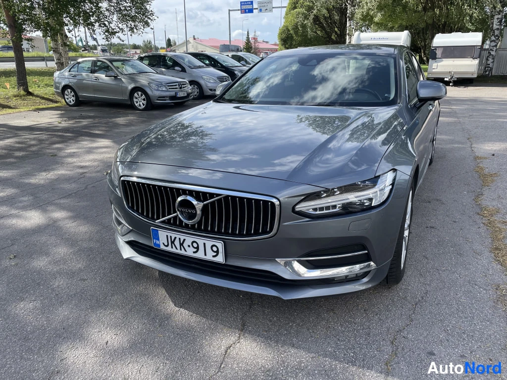 Etuviisto, nokka oikealle: Volvo S90:n etunäkymä, rekisterikilpi, tummassa parkkipaikassa muina autoina taustalla.