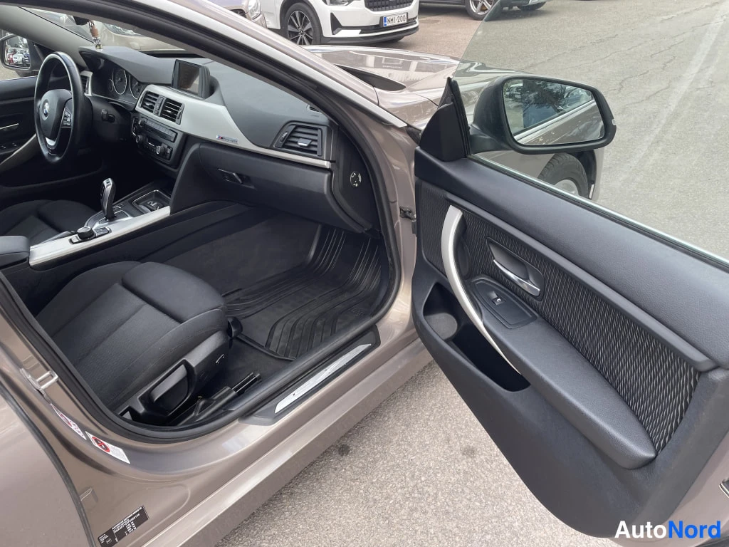 Sivulta: BMW:n etupenkki ja kojelauta avoimesta etuovesta kuvattuna, tumma matto ja kangasistuin näkyvät.