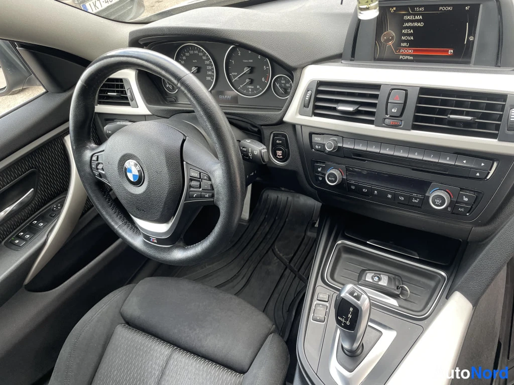 Edestä: BMW:n ohjaamo: ratin BMX-tunnus, kojelaudan analogiset mittarit ja infotainment-näyttö, jonka radiolista näkyy (ISKELMA, JÄRVIRADIO, KESKINADIO).