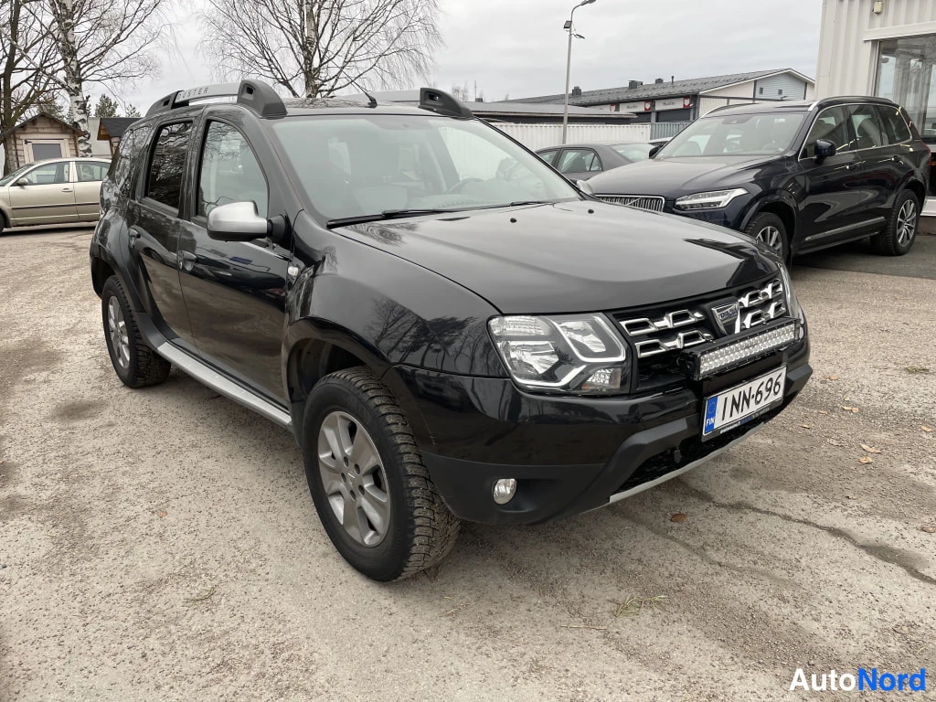 Etuviisto, nokka oikealle: Musta Dacia Duster etuviistosta oikealta, etupanssariin asennettu lisävalo ja kattokaiteet näkyvät.