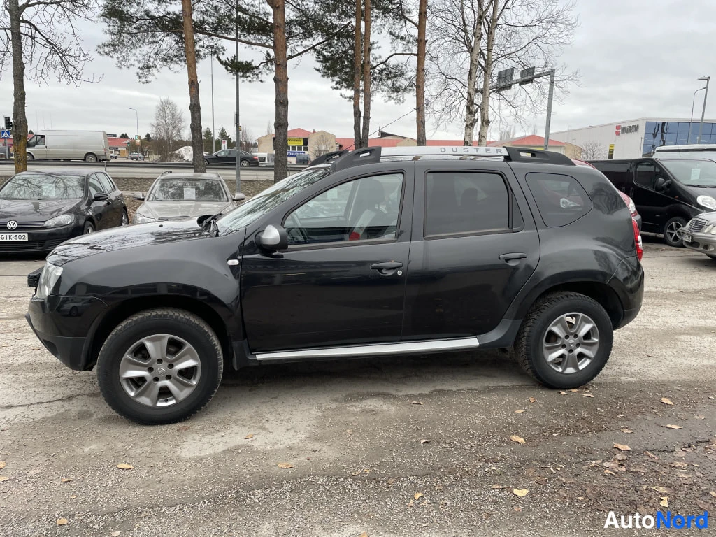 Sivulta: Mustan Dacian sivukuva autopaikalla, kattokaiteet ja ovien alaosien kromilistat näkyvissä.