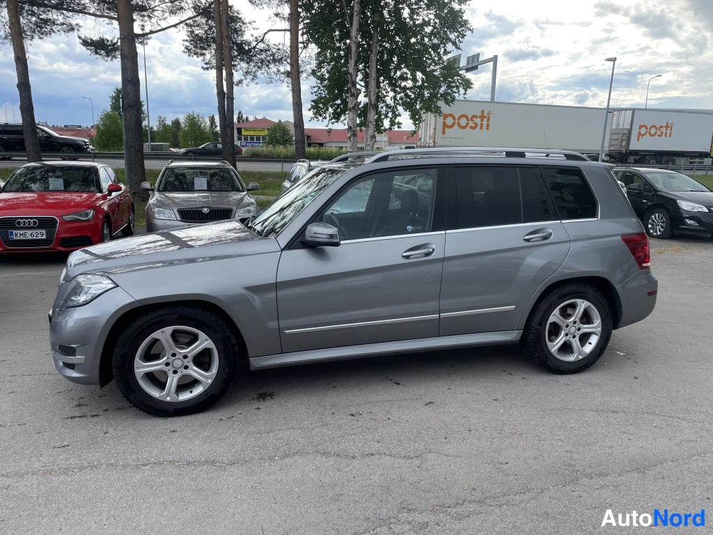 Etuviisto, nokka vasemmalle: Mercedes-Benz GLK:n etukulma vasemmalta: hopeaharmaa runko, kattokaiteet ja hopeavanteet näkyvät tiili vaihtoautoparkin taustalla.