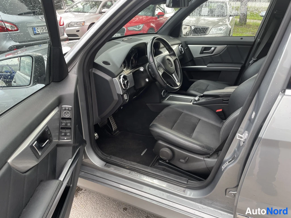 Muu: Mercedes-Benz GLK:n etumatkustamo vasemmasta ovesta katsottuna: tumma sisusta, Mercedes-ohjauspyörä, ikkunanokeimet ovelassa.