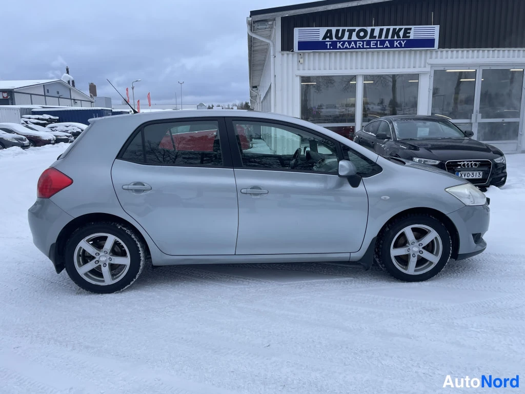 Etuviisto, nokka oikealle: Toyota Auris -henkilöauto sivulta, hopeinen koripinta, viisiovinen viistoperä, lumisella pihalla autoliikkeen edessä.