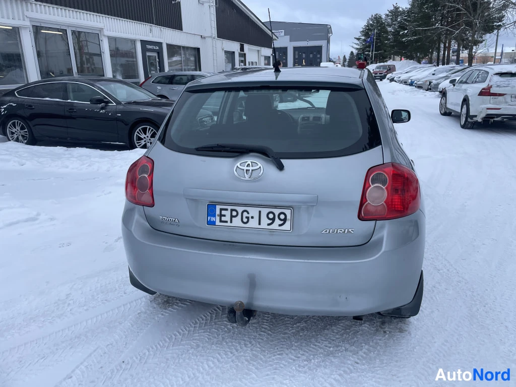 Takaa: Toyota Auris -henkilöauto suoraan takaa, hopeinen koripinta, rekisterikilpi, takavalojen välissä Toyota-merkki, lumisella pihalla.