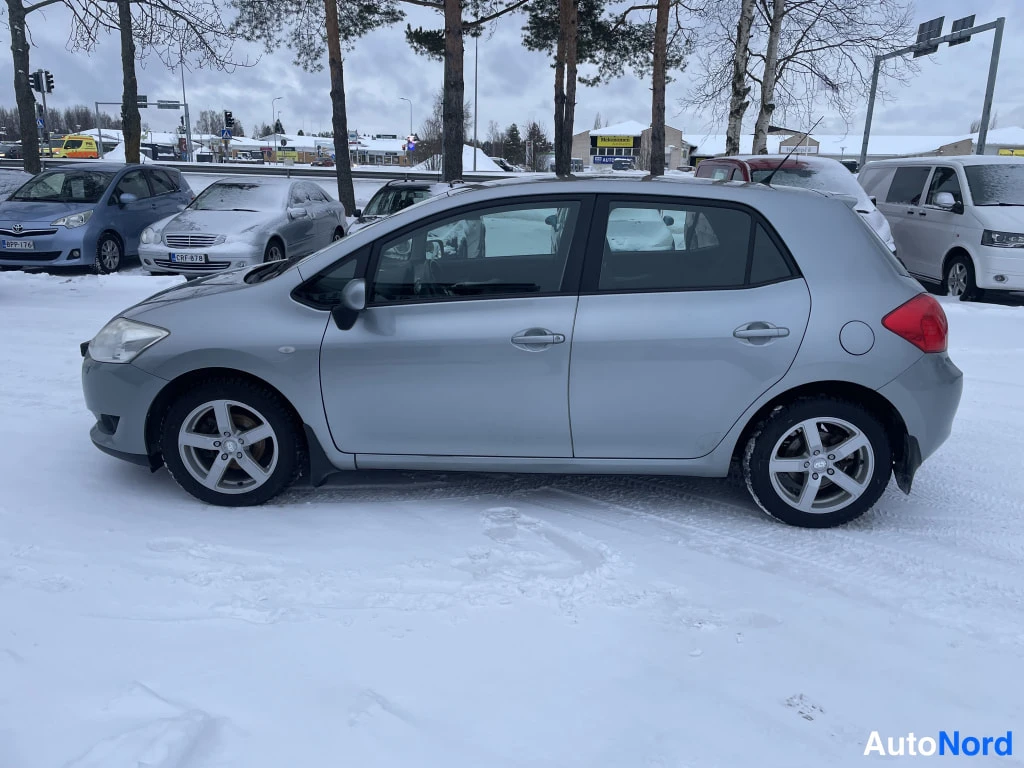 Etuviisto, nokka vasemmalle: Toyota Auris -henkilöauto kulmassa etupää kuvassa vasemmalla, hopeinen koripinta, viisiovinen viistoperä, lumisella parkkipaikalla.