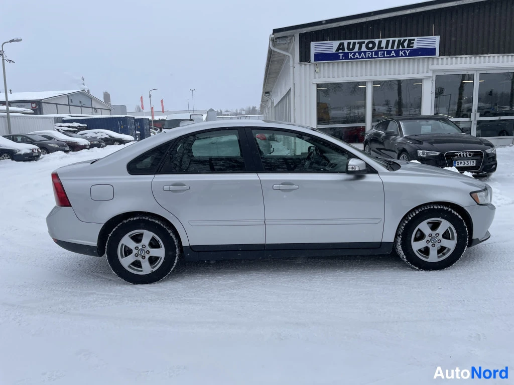 Sivulta: Hopeinen sedan sivusta vasemmalta lumisella paikalla, autoliikkeen rakennus taustalla.