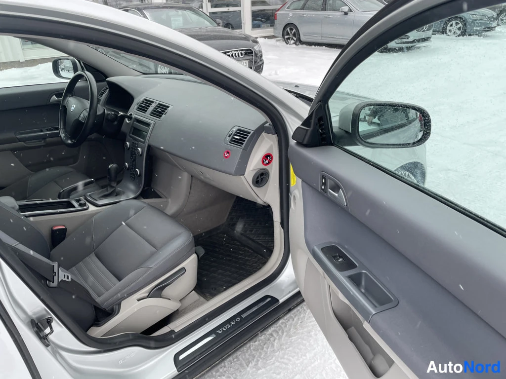 Sivulta: Kuljettajan puolen matkustamo avoimesta ovesta: Volvo-ohjauspyörä, tumma kojelauta ja ovipaneeli, lunta ulkona.