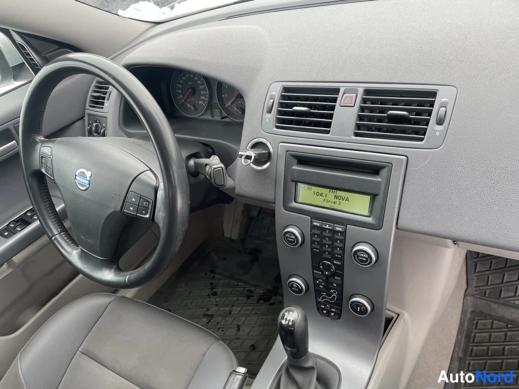Sivulta: Kojelauta lähikuvassa: Volvo-ohjauspyörä, mittaristo, radiopaneeli ja näytöllä teksti '104.1 NOVA'; käsivaihteisto.
