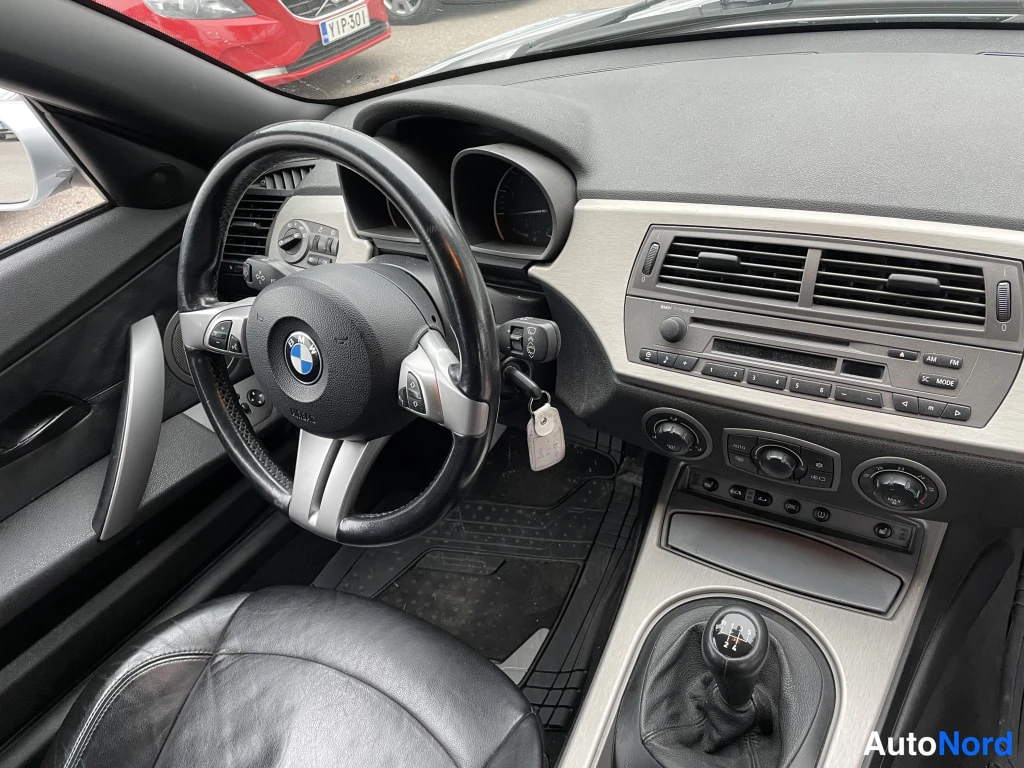 Edestä: Ohjaamo lähikuvassa: BMW-logo ohjauspyörässä, manuaali vaihdekeila ja kojelaudan ilmastointi- ja radiopainikkeet.