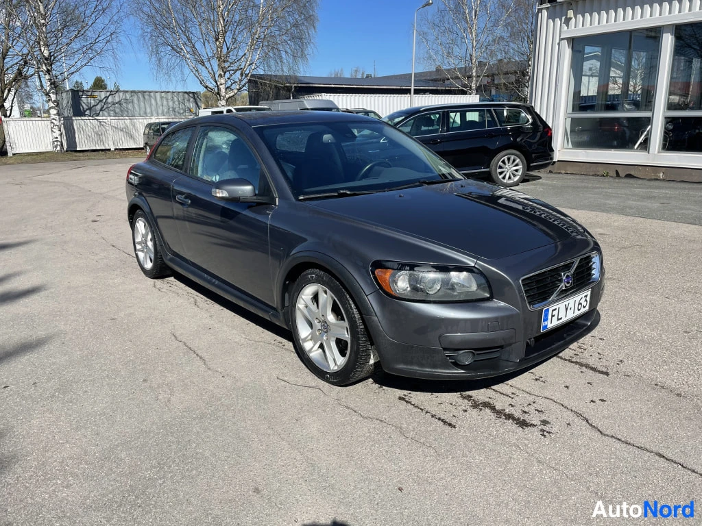 Etuviisto, nokka oikealle: Tummanharmaa kolmiovinen henkilöauto etukulmasta oikealta, rekisterikilpi.