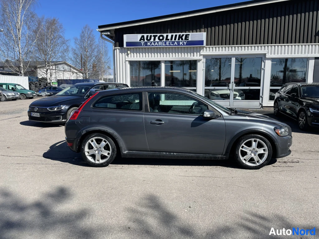 Etuviisto, nokka oikealle: Tummanharmaa kaksioinen hatchback sivulta etuosaa vasten kuvattuna autoliikkeen pihalla.