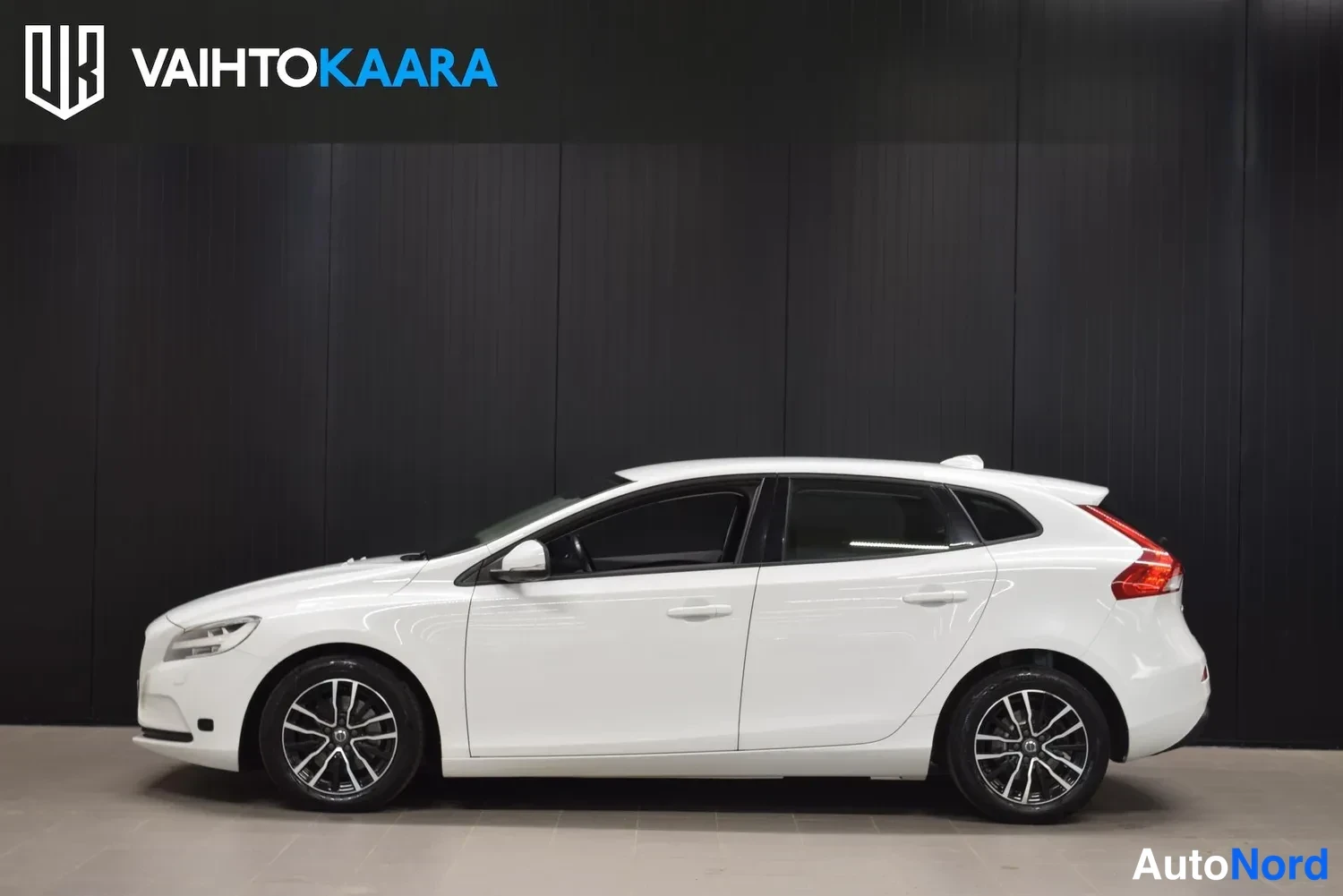 Etuviisto, nokka vasemmalle: Valkoinen viisiovinen hatchback sivulta kuvattuna, keula vasemmalla, tumma tausta ja vaalea lattia.