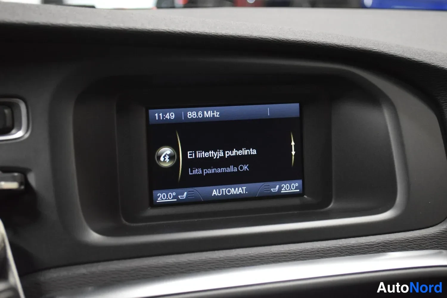 Yksityiskohta: Auton kojelaudan infotainment-näyttö näyttää kelloa 11:49, radioasemaa 88.6 MHz ja tekstiä 'Ei liitettyjä puhelinta – Liitä painamalla OK', alareunassa lämpötil
