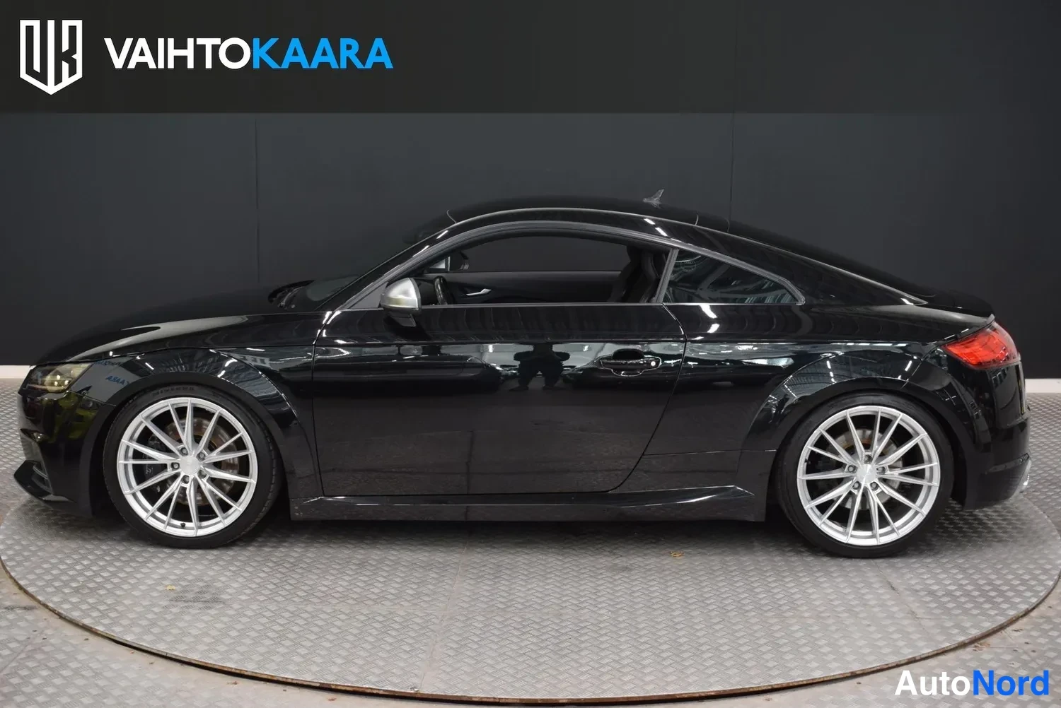 Sivulta: Mustan Audi TT-coupeen kylkinäkymä molempien ovien ja pyörien osalta studio-alustalla, taustana tumma seinä ja Vaihtokaara-logo.