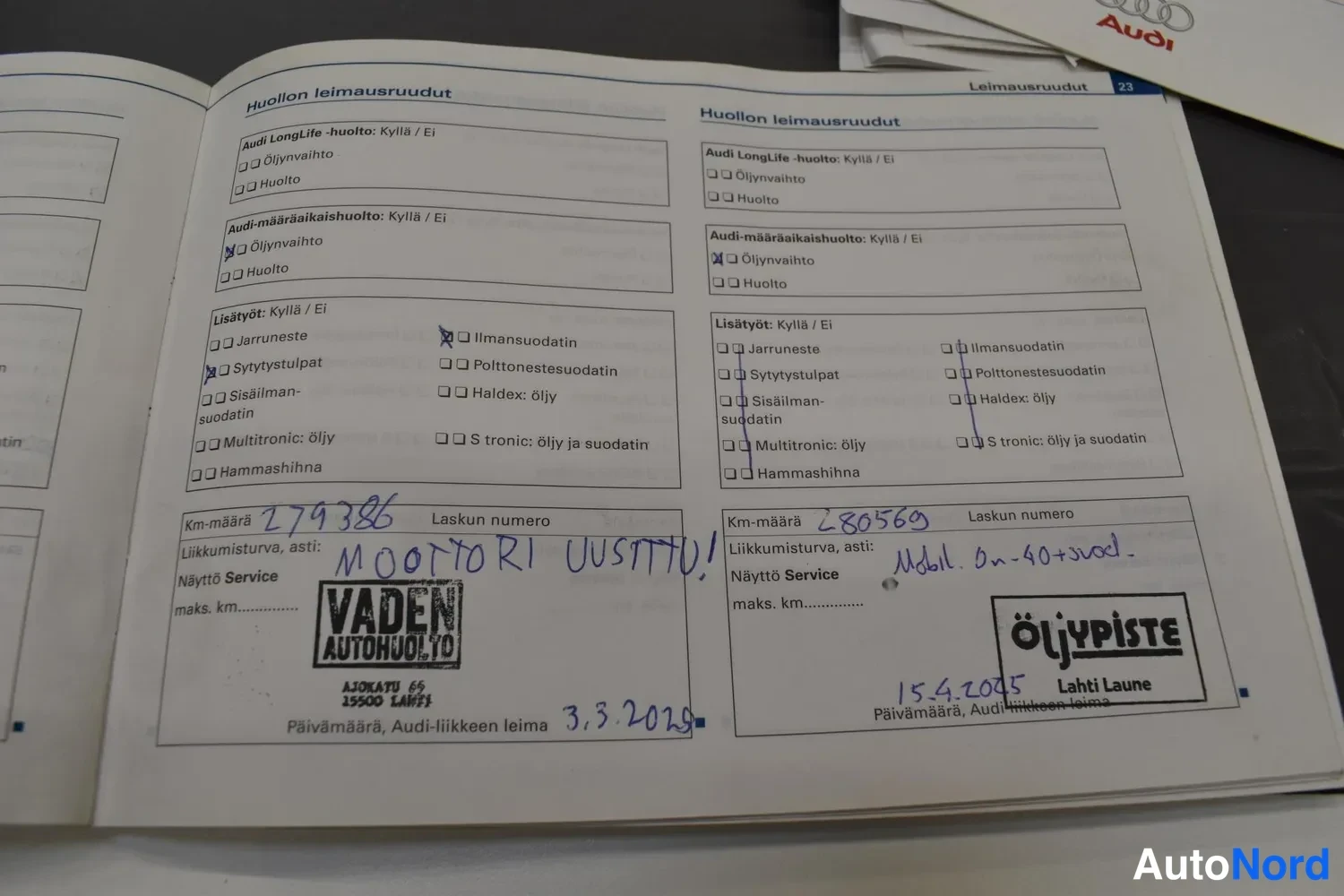 Muu: Audi-huoltokirjan aukeama kahdella huoltoleimalla: vasemmalla Vaden Autohuollon leima 3.3.2020 ja km-määrä 279386, oikealla Öljypisteen leima 15.4.2025 ja km-mä