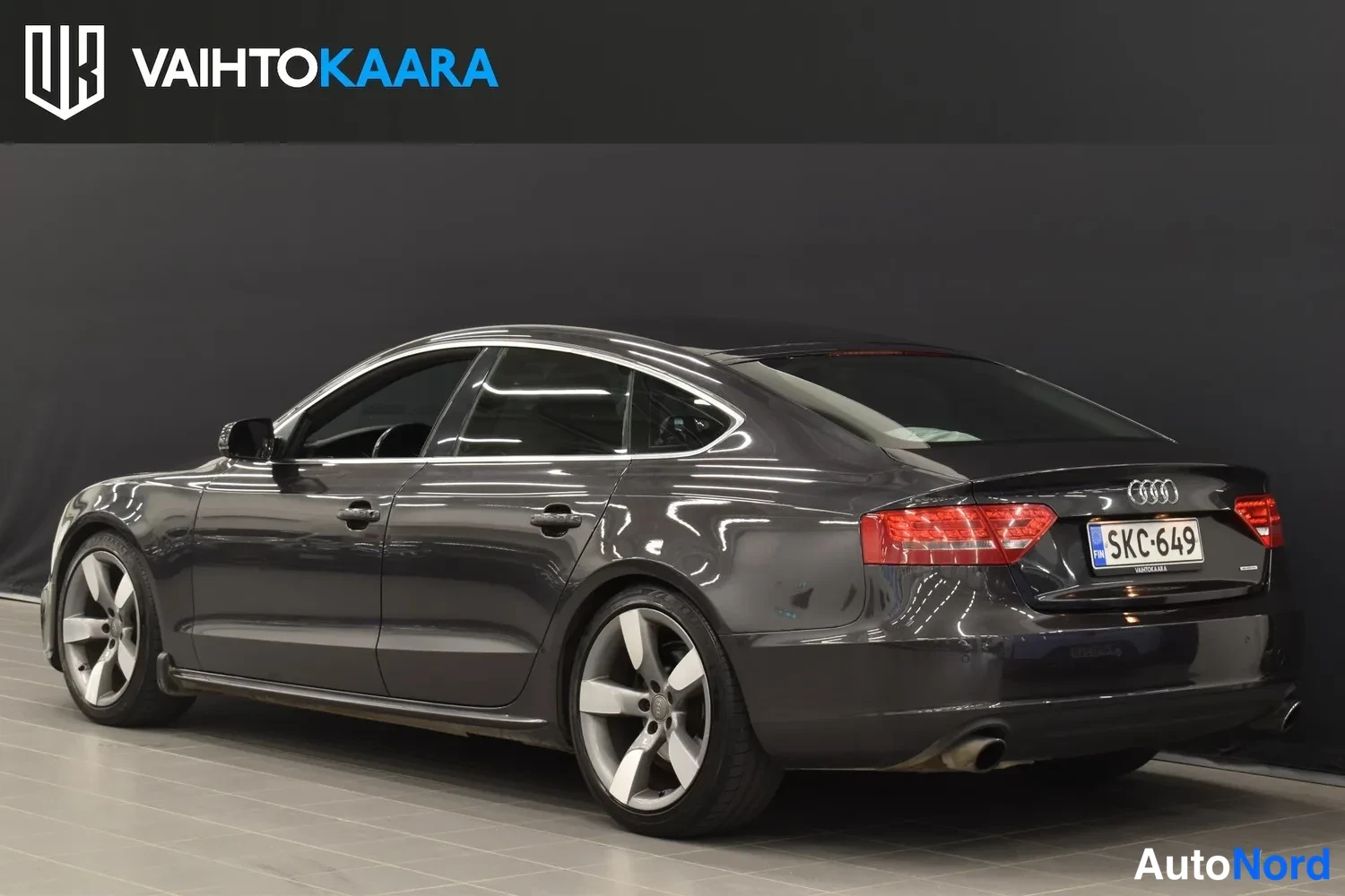 Takaviisto: Audi A5 Sportback takapäältä oikealta, tummanharmaa kori, punaiset takavalot, rekisteri.