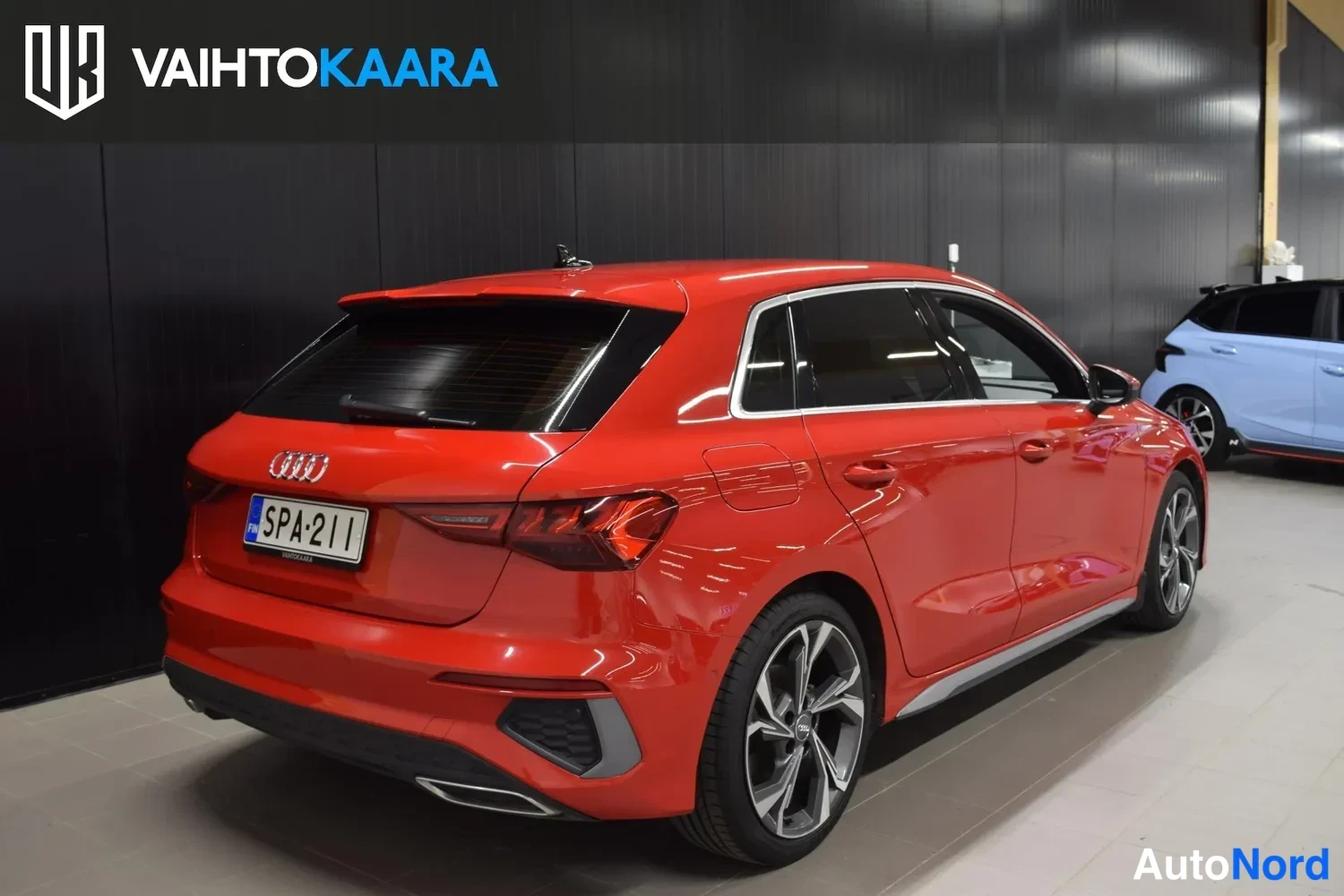 Takaviisto: Punaisen Audi A3 Sportback -auton takanurkkakuva sisätiloissa; takavalot, tavaratilan luukku, Audi-merkki ja rekisterikilpi näkyvät.