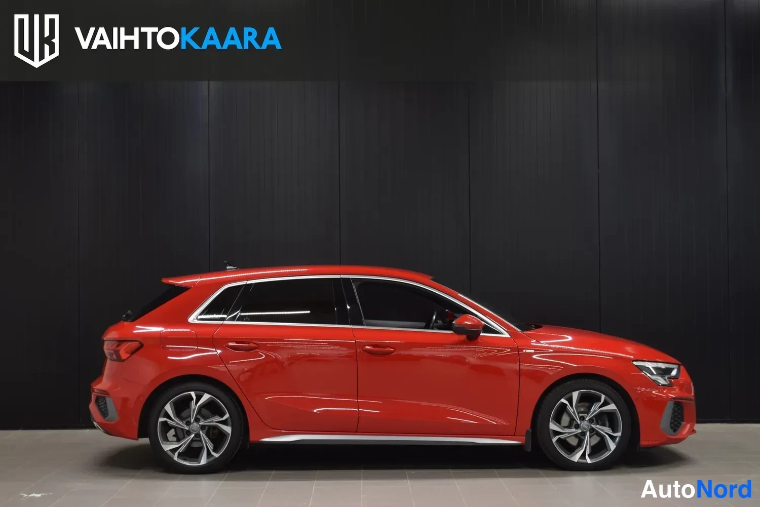 Sivulta: Punaisen Audi A3 Sportback -auton sivukuva; kattolinja, ovet ja sektorivanteet näkyvät tummaa seinää vasten.