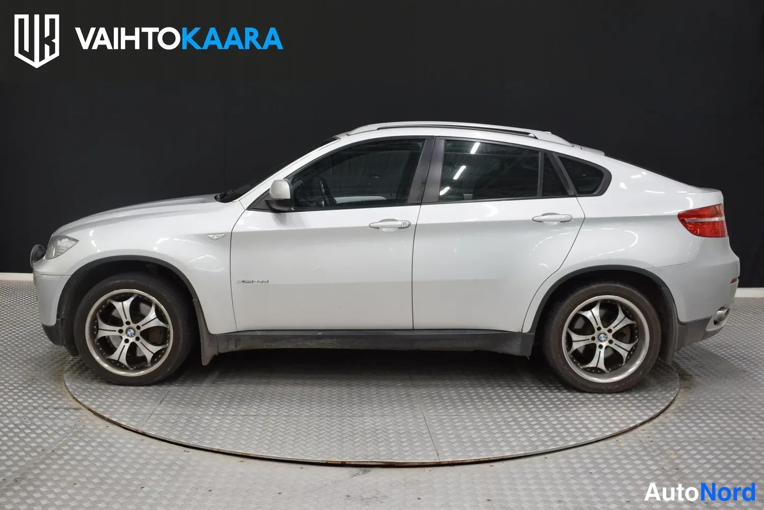 Sivulta: Hopeanvärinen BMW X6 kyljestään vasemmalta kuvattuna sisätiloissa; kattokaiteet ja monipolttoiset kevytmetallivanteet näkyvissä.