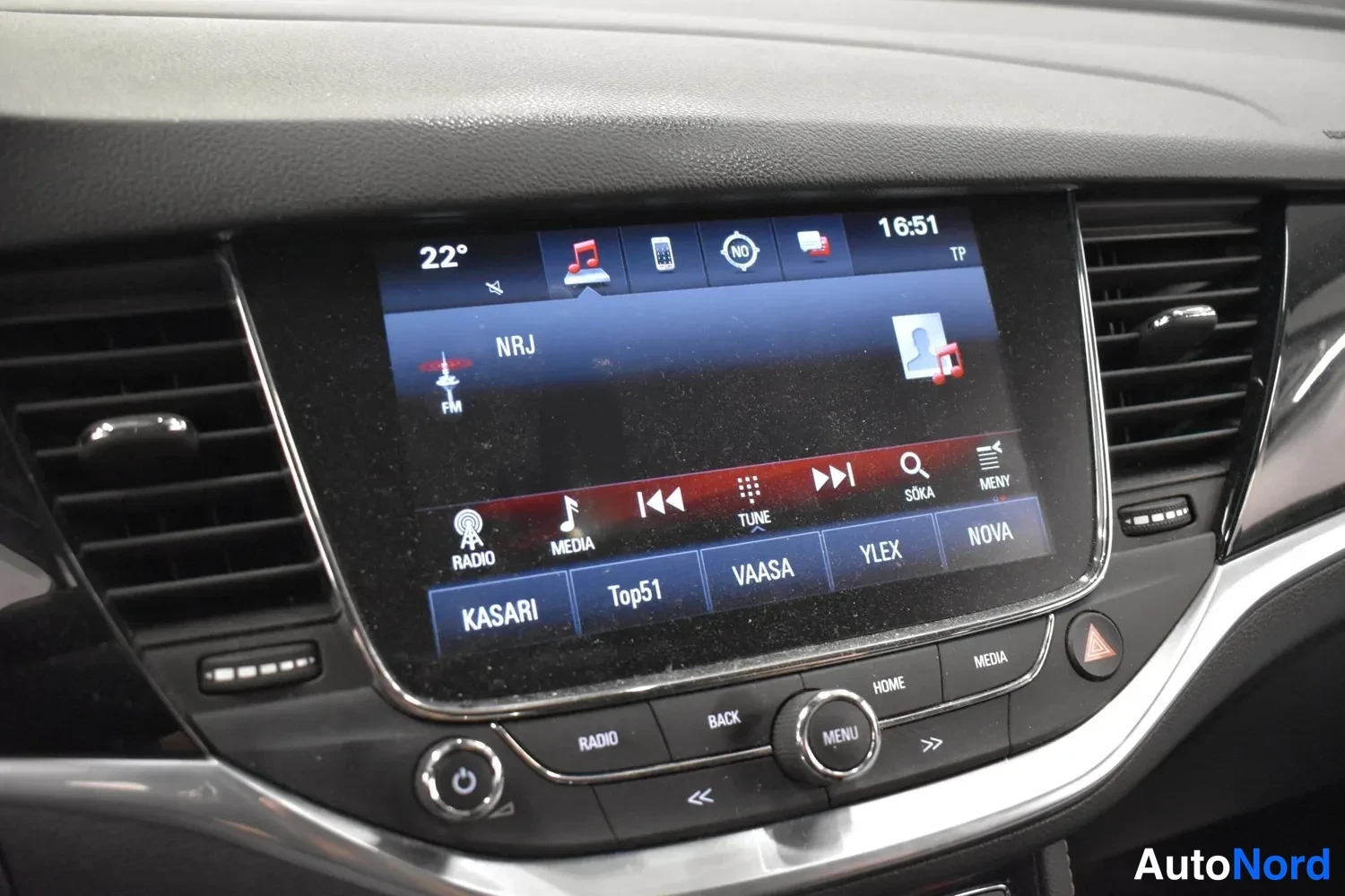 Muu: Keskikonsolan infotainment-näyttö näyttää radiota (NRJ, FM), lämpötilan 22° ja kellon 16:51; alla RADIO-, BACK- ja MENU-napit.