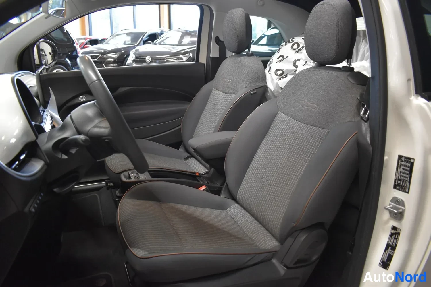 Yksityiskohta: Fiat 500:n sisätilat: ohjauspyörä vasemmalla, kaksi harmaata kangasistuinta oranssein reunoituksin ja keskin päällystetty varusteilla.