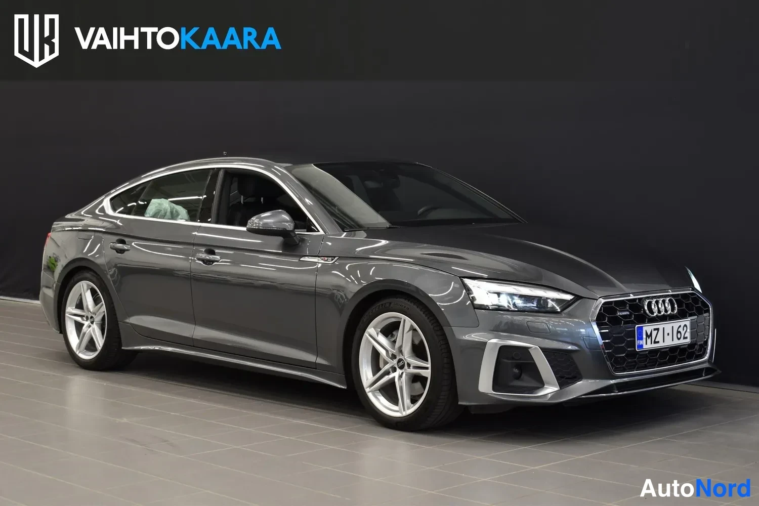 Etuviisto, nokka oikealle: Audi A5 Sportback etuosoalta oikealta kuvattuna, tummanharmaa kori, hopeavanteet ja rekisteritunnus.