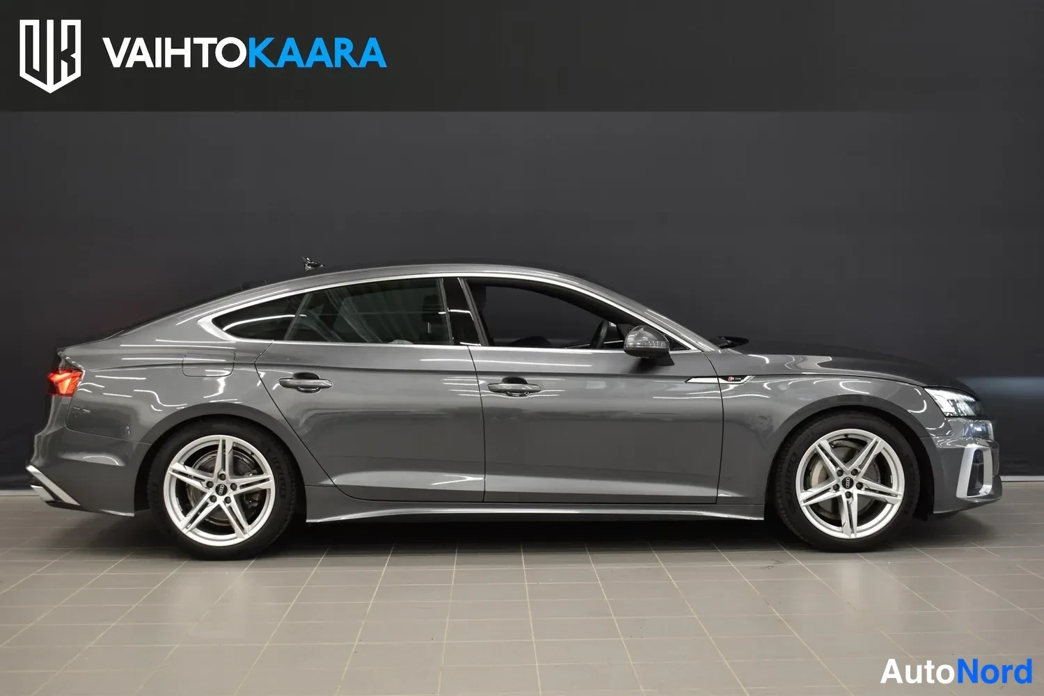 Sivulta: Harmaan Audi A5 Sportbackin sivukuva sisätiloissa, koko oikea sivu ja molemmat pyörät näkyvät.
