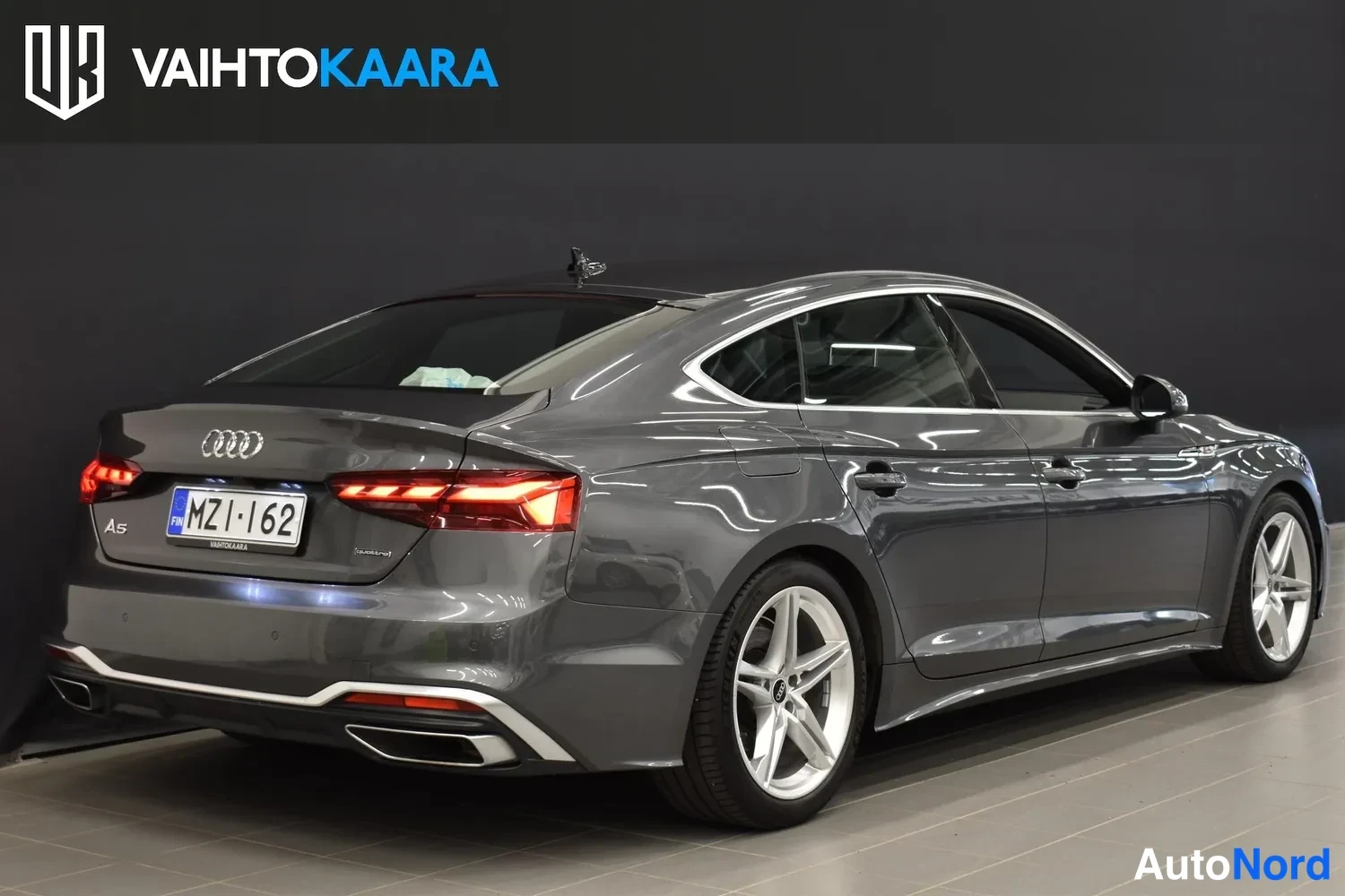Takaviisto: Harmaan Audi A5 Sportbackin takavino kuva sisätiloissa, takavalot, perä ja rekisteritunnus näkyvät.